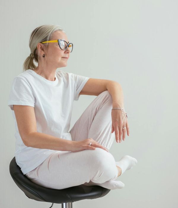 A woman in a calm, seated yoga pose against a soft mauve background.